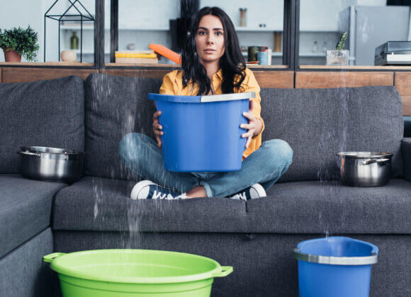 Brunette woman holding bucket on sofa during water damage in living room