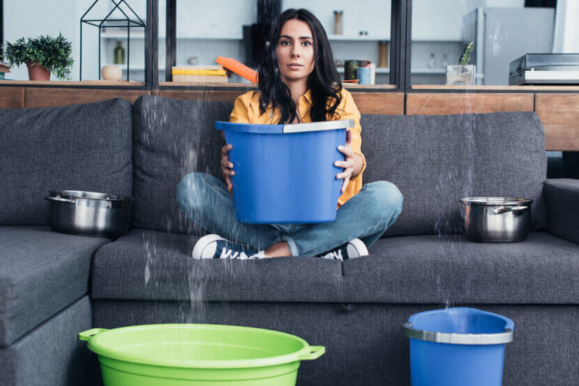 Brunette woman holding bucket on sofa during water damage in living room