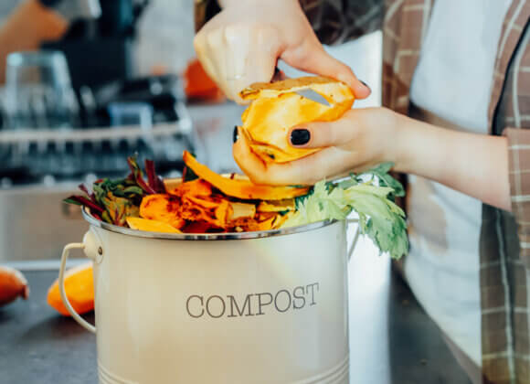 Kompostieren in der Wohnung: Funktioniert das wirklich? Compost the kitchen waste, recycling at home. Compost bin with vegetables cutted leftovers on the kitchen table with blurred woman cooking meal. Environmentally responsible, ecology. Selective focus