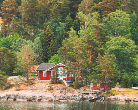 Sweden. Beautiful Red ASwedish Wooden Log Cabins Houses On Rocky Island Coast In Summer. Lake Or River Landscape